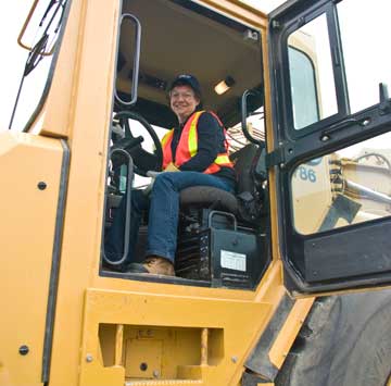 women sitting in cab of earth moving machine, smiling down at the photographer; she is wearing a hard hat and red/yellow vest